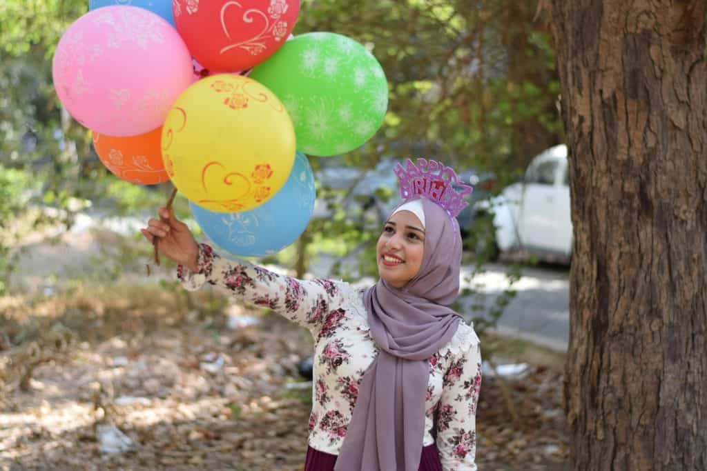 A Muslim woman, wearing hijab, with a birthday hat and holding balloons
