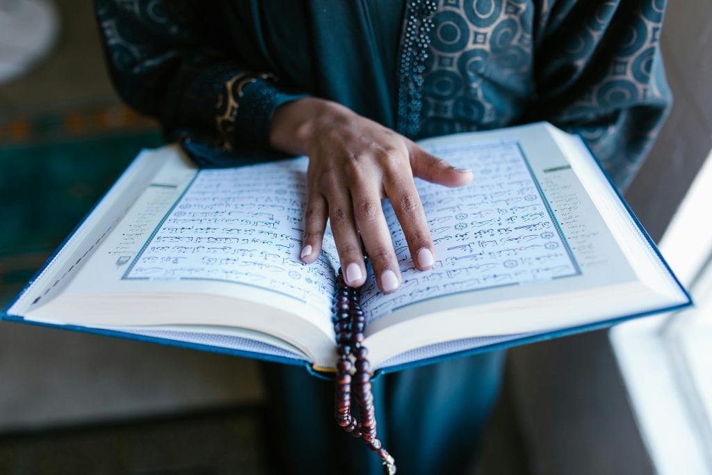 A person in a blue thobe holding a Quran, representing praying in Arabic.